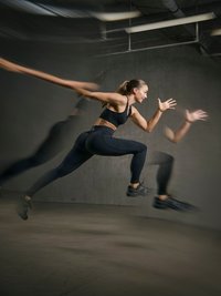 Γυναίκα με αθλητικό μαύρο ντύσιμο που τρέχει γρήγορα σε εσωτερικό χώρο με θόλωμα κίνησης, μπροστά από γκρι τσιμεντένιο τοίχο.