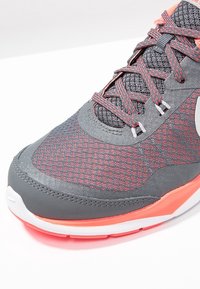 Harmaa urheilukenkä teksturoidulla päällisellä, kontrastivärisillä punaisilla yksityiskohdilla ja valkoisilla yksityiskohdilla pohjassa. Siinä on pyöreät nauhat ja pehmustettu kaulus.