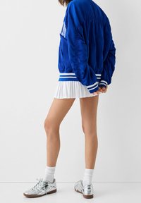 En person iklädd en blå bomberjacka, vit plisserad minikjol, vita strumpor och metalliska silverfärgade sneakers som står med händerna knäppta bakom ryggen.