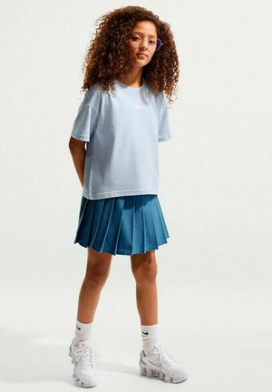 Persona joven con cabello rizado que lleva gafas, camiseta Nike azul claro, falda plisada azul, calcetines blancos Nike y zapatillas blancas Nike, de pie.