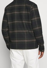 Muž v tmavě zelené a hnědé kostkované košili s dlouhým rukávem a bílých kalhotách, zobrazený zezadu proti jednobarevnému pozadí.