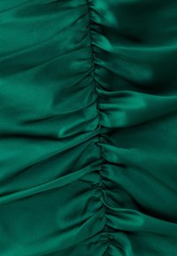 Satinmaterial i djup grön med diagonala rynkdetaljer. Slät textur och lyster, som framhäver mjuka veck och samlingar genom hela tyget.