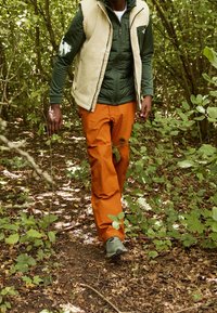 Persona che cammina lungo un sentiero nel bosco indossando una giacca verde oliva, un gilet beige, pantaloni arancioni e scarpe da trekking grigio chiaro.