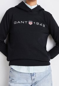 Sweat à capuche noir avec le logo "GANT 1949", comportant un texte blanc et un graphisme de bouclier. Tissu doux, coupe ajustée, associé à une chemise à rayures bleues.
