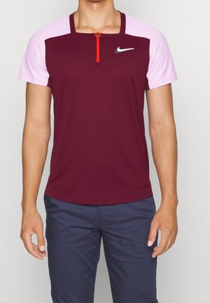 Homme portant un t-shirt Nike manches courtes bordeaux et rose avec une fermeture éclair à l'avant et un pantalon bleu foncé, debout les bras détendus le long du corps.