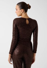 Tuta marrone con paillettes e maniche lunghe. Presenta un'apertura a goccia sulla schiena e un design aderente, con una superficie testurizzata.