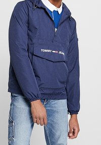 Marinblå tröja med huva, elastiska ärmslut, framfickor och "TOMMY JEANS"-logotyp. Tillverkad av ett lätt och slätt syntetiskt material.