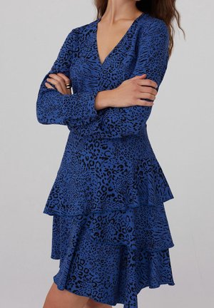 Kobieta ubrana w niebieską sukienkę z długim rękawem i dekoltem w serek, z czarnym nadrukiem w panterkę i warstwowym falbanowym dołem, z założonymi skrzyżowanymi rękami na piersiach.