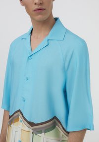Camicia a maniche corte realizzata in tessuto azzurro chiaro, con una stampa grafica colorata lungo il fondo che ricorda forme architettoniche.