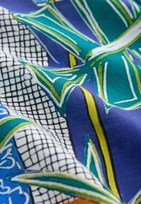 Tissu en coton avec un motif de feuilles bleu et vert, surmonté d'accents de grille noirs. Texture douce avec des couleurs vives et des détails complexes.