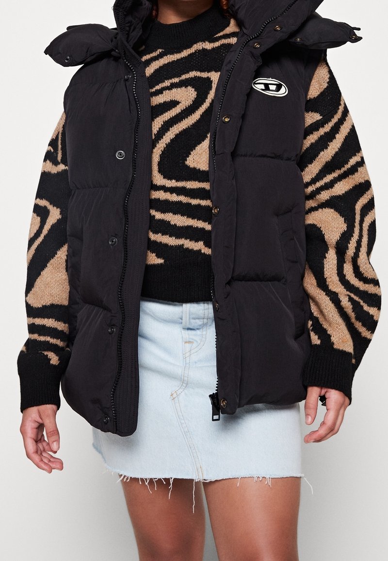 Svart vadderad väst med hög krage, med dragkedja och knappstängning, bärs över en svart och beige mönstrad tröja och en jeanstub.