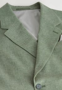 Blazer vert texturé avec un col cranté, mettant en valeur une doublure grise claire et une fermeture à un bouton. Les détails incluent un subtil motif en chevrons.