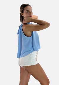Top senza maniche di un blu chiaro con un collo arricciato, abbinato a pantaloni corti in denim bianco strappato. Lo sfondo è bianco semplice.