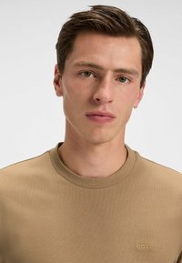 Light brown crew-neck t-shirt made of smooth fabric, featuring subtle ribbed texture and a small embroidered logo on the chest.