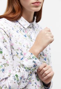 Woman wearing white floral long-sleeve shirt fastening the cuff with her right hand, visible from shoulders to lips against a white background.