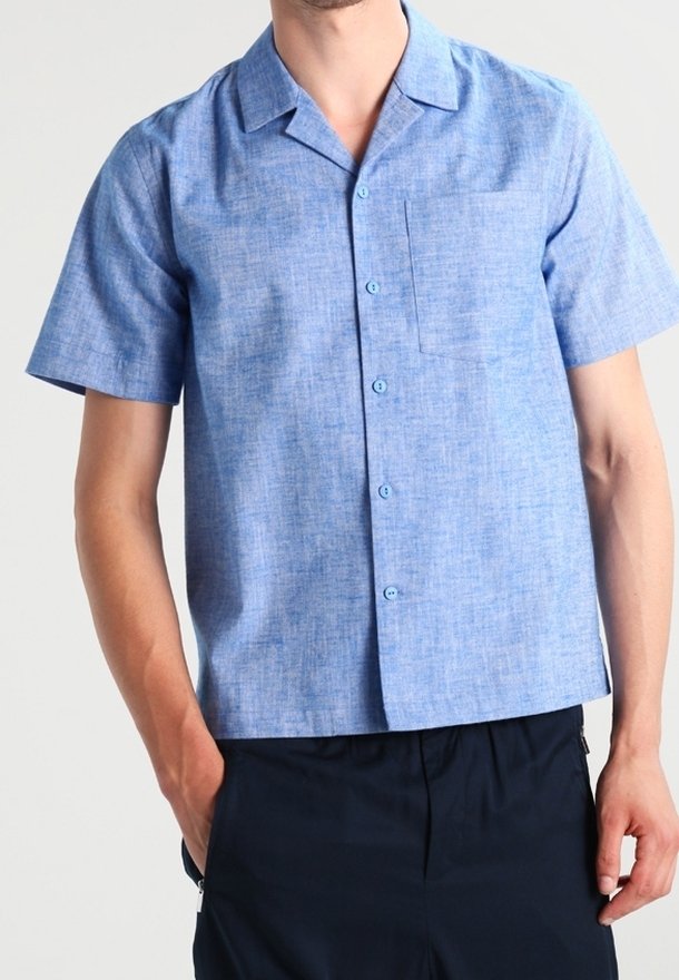 Man som bär en ljusblå kortärmad skjorta med knappar och bröstficka samt mörka byxor, med händerna vilande i fickorna.