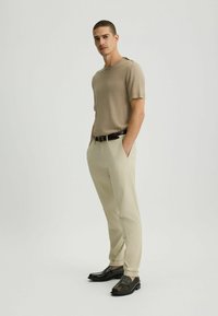 Camiseta de punto beige de manga corta, chinos crema, cinturón de piel negra, zapatos negros elegantes, pose de pie con las manos en los bolsillos.