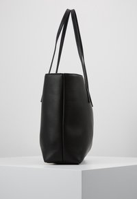 Bolso de tote negro de cuero texturizado con largas correas dobles para el hombro, mostrado de pie sobre un pedestal blanco contra un fondo liso.