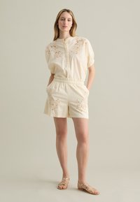 Conjunto de algodón color crema que incluye una blusa de manga corta con detalles bordados y pantalones cortos con cintura elástica, combinados con sandalias de tiras.