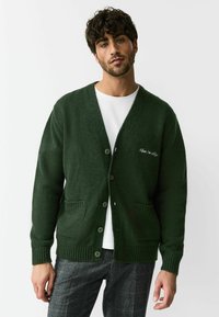 Donkergroene gebreide cardigan met een V-hals, vier knopen, twee voorzakken en geribbelde manchetten. Heeft een witte tekstborduring op de linkerborst.