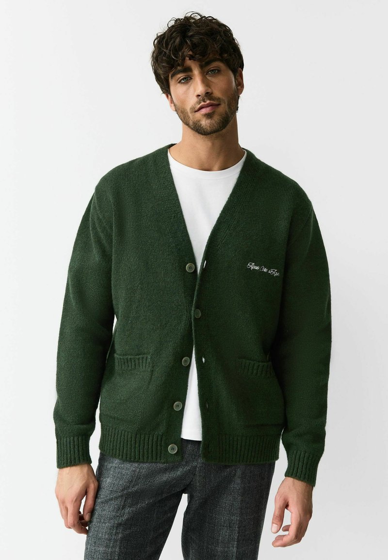 Donkergroene gebreide cardigan met een V-hals, vier knopen, twee voorzakken en geribbelde manchetten. Heeft een witte tekstborduring op de linkerborst.