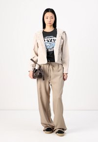 Veste en velours côtelé beige courte, t-shirt noir avec graphique, pantalon ample beige, petit sac à main marron, et chaussures noires avec semelles beiges.