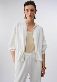 Femme portant un sweat à capuche blanc zippé et un pantalon assorti sur un débardeur beige, accessoirisée avec des bracelets en or et des boucles d’oreilles triangulaires.