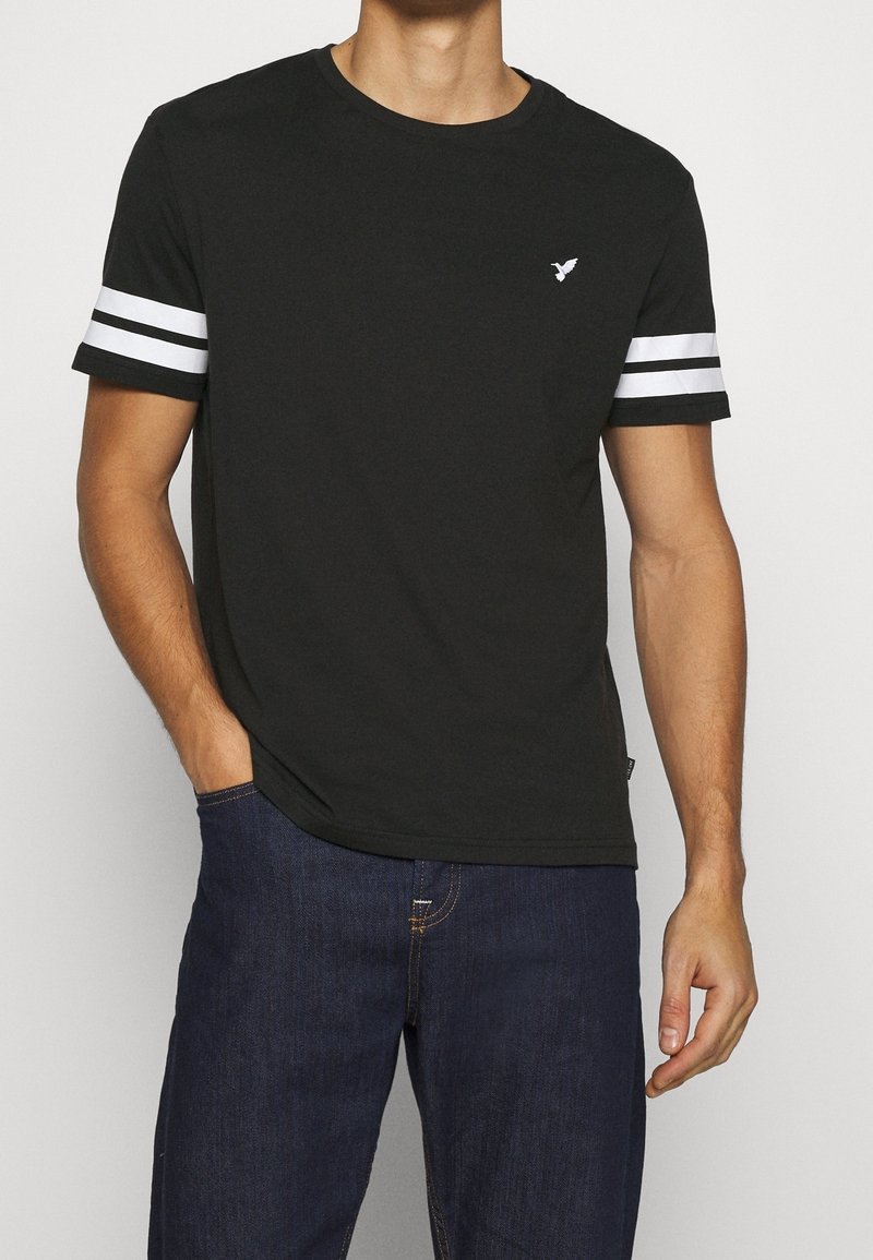 T-shirt en coton noir avec des accents de rayures blanches sur les manches courtes et un petit logo sur la poitrine. Encolure ronde classique, coupe décontractée.