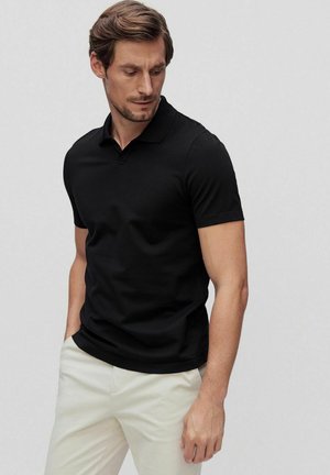 Black short-sleeve polo shirt with a classic collar, smooth texture, and a fitted design, paired with light-colored trousers.