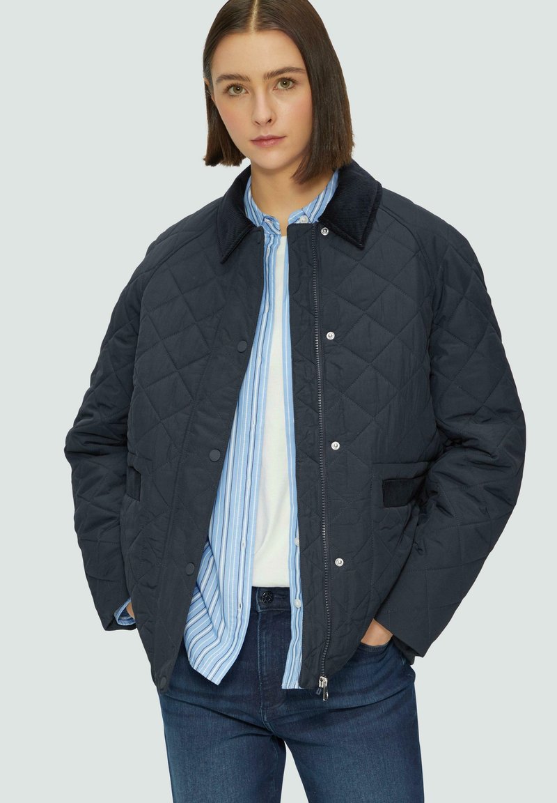 Veste matelassée bleu marine avec un col en velours côtelé, boutons-pression, poches latérales et un motif en losange. Portée sur une chemise rayée.