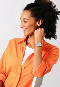 Silver-tonad armbandsur med en rund urtavla, metallarmband och texturerade detaljer. Buret på en handled mot en bakgrund av en orange skjorta.