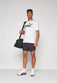 Valkoinen lyhythihainen t-paita, jossa PUMA-logo, mustat urheilushortsit, musta olkalaukku ja valkoiset kengät punaisilla yksityiskohdilla. Neutraali tausta.