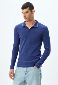 Marinblå långärmad polo med ribbad textur, kontrasterande randig krage och en avslappnad passform. Bärs med ljusblå jeans.