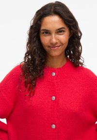 Femme aux longs cheveux bouclés portant un cardigan moelleux rouge vif avec des boutons en métal argenté, souriant doucement devant un fond blanc uni.