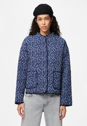 Navy quilted jacket with small white flower patterns, round neckline, front zip, and two large patch pockets. Soft texture and relaxed fit.
