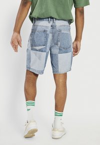 Denim shorts featuring a patchwork design in light and dark blue tones, with two back pockets and a frayed hem. Worn with white socks and sneakers.