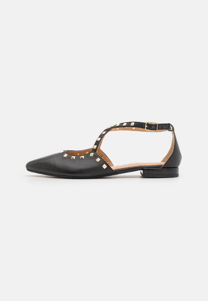Chaussure plate noire à bout pointu avec bride à la cheville, ornée de clous carrés dorés et d'une petite boucle sur un fond blanc uni.