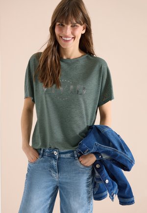 Smilende kvinde iført grøn T-shirt og lyseblå jeans, der holder en blå denimjakke i venstre hånd mod en beige baggrund.