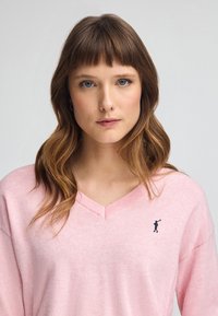 Maglione rosa chiaro a scollo a V realizzato in un tessuto morbido, con dettagli a coste e un piccolo logo a contrasto sul lato sinistro del petto.