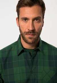 Camisa de cuadros verde con botones y cuello, que presenta patrones de cuadros en tonos más oscuros y más claros, un bolsillo en el pecho y botones oscuros.