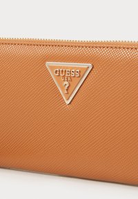 Brown textured wallet featuring a triangular logo plate with "GUESS USA" text and a question mark, secured with a gold zipper.