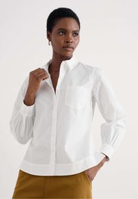 Camicia bianca con colletto e maniche lunghe a sbuffo, con una leggera trama e taschino sul petto. Abbinata a pantaloni beige.