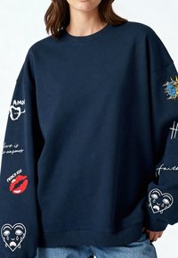 Felpa oversized blu navy in cotone, con design ricamati sulle maniche, tra cui cuori, labbra e testi come "Bacio Francese."