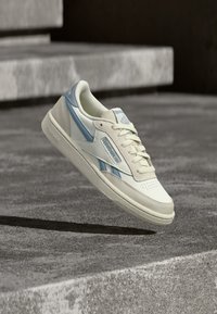 Bijela i svijetloplava Reebok tenisica koja leti iznad teksturirane betonske površine s betonskim stepenicama u pozadini.