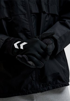 Person mit schwarzen Handschuhen mit weißen Chevron-Symbolen, der den Handschuh an der anderen Hand über einer schwarzen Reißverschlussjacke und grauen Hosen anpasst.