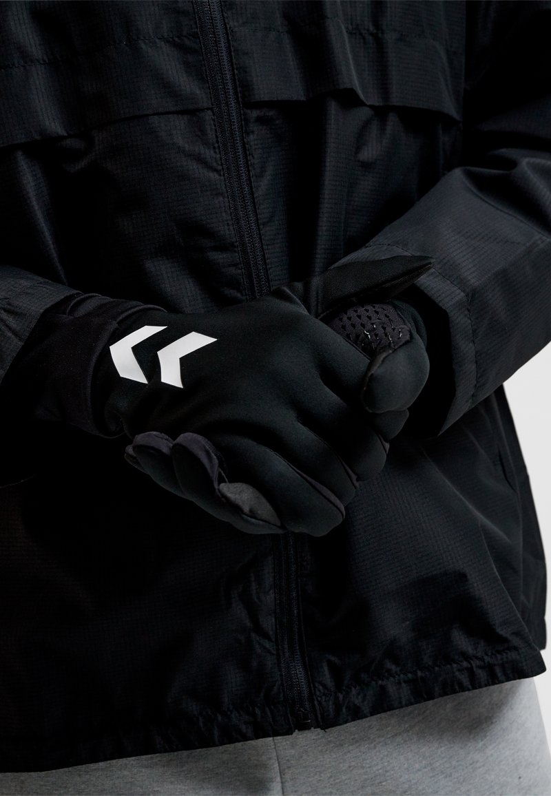 Personne portant des gants noirs avec des symboles en chevron blancs, ajustant un gant sur l'autre main par-dessus une veste noire zippée et un pantalon gris.