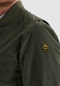 Mann trägt eine dunkelgrüne Jacke mit geripptem Kragen und PME Legend American Classic Aufnäher am oberen Ärmel.