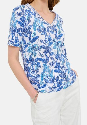 Femme portant un t-shirt blanc à manches courtes et col en V avec des motifs floraux et feuillus bleus, associé à un pantalon blanc et un bracelet en argent.