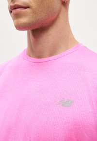 Κοντινό πλάνο ενός ατόμου που φοράει ένα ροζ διάτρητο αθλητικό μπλουζάκι με ανακλαστικό λογότυπο New Balance στο στήθος.