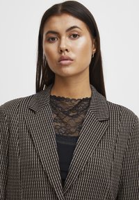 Černobílý pruhovaný blazer s špičatými klopami, vrstvený přes černý krajkový top s květinovými vzory. Hladká textura, přiléhavý střih.